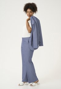 Completo su misura azzurro chiaro con pantaloni a gamba larga, tessuto strutturato e una giacca appoggiata sulla spalla. Il modello indossa una maglietta bianca e sandali con tacco.