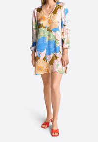 Nainen, jolla on päällään värikäs kukallinen V-pääntie mekko, jossa on pitkät hihat, ja punaiset avokärkiset korot, yksinkertaista valkoista taustaa vasten.