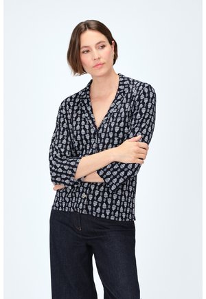 Camisa de manga larga con cuello en color azul marino y patrón de hojas blancas, confeccionada en tela ligera, con detalle de botones en la cintura, combinada con jeans oscuros.