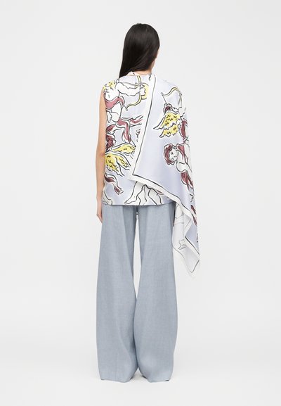 Γυναίκα με μακριά σκούρα μαλλιά φοράει ανοιχτόχρωμο μπλε παντελόνι φαρδύ και αμάνικο τοπ με ένα λευκό φουλάρι που έχει σχέδια με αγγελάκια και φτερά.