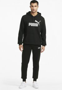 Ensemble de sweat à capuche noir et de pantalons de survêtement en tissu doux, avec un grand logo Puma blanc sur la poitrine et la jambe gauche du pantalon. Des baskets blanches complètent le look.