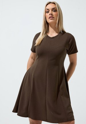 Kobieta z długimi blond włosami, ubrana w brązową sukienkę z krótkim rękawem do kolan z kieszeniami, stojąca na tle jednolitego jasnego tła.