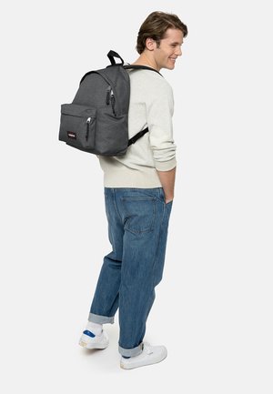 Mochila de tela gris con un bolsillo frontal, cremalleras negras y correas ajustables, llevada sobre un suéter ligero y combinada con pantalones de mezclilla relajados.