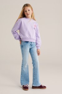 Noor tüdruk seisab käsi puusal, kandes lavendlivärvi lillemustriga dressipluusi, helesiniseid teksaseid ja burgundia värvi kingi.