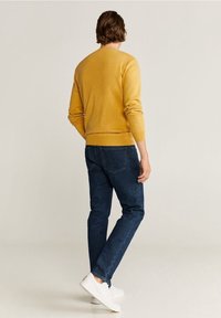 Muž stojící zády, oblečený v hořčicově žlutém svetru, tmavě modrých džínách a bílých teniskách, opřený o jednobarevné světlé pozadí.