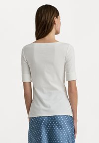 Vit, åtsittande bomullströja med rundad halsringning och uppvikta ärmar, kombinerad med en blå prickig kjol. Slät textur, enkel design.