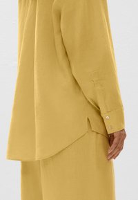 Chemise en lin jaune à manches longues, ourlet arrondi et fentes sur les côtés. Détail de bouton au niveau du poignet montrant une texture lisse et une coupe décontractée.