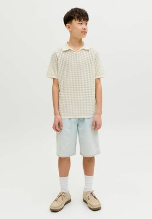 Jauns cilvēks stāv, valkā bēša rakstainu īsroku krekliņu, gaišas džinsa bikses līdz ceļiem, baltas zeķes un bēšas auklu apavus.