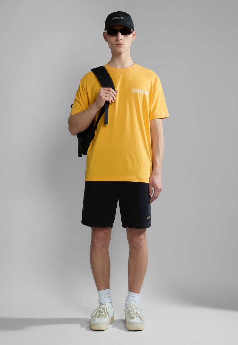 Gele katoenen t-shirt met logo, zwarte shorts, witte sokken en beige sneakers. Zwarte pet en zonnebril, staand tegen een grijze achtergrond.