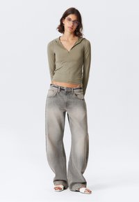 Olive green long-sleeve crop top with zip detail, paired with light gray wide-leg jeans featuring fading and subtle stitching, completed with white sandals.