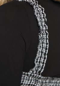 Svart topp med volangiga svart-och-vita rutiga axelremmar; texturerat tyg med kontrasterande mönster. Närbild av axelområdet.