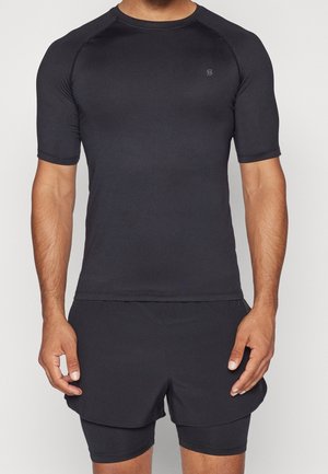 Atletische man met een aansluitend zwart T-shirt met korte mouwen en gelaagde zwarte hardloopshorts, staand tegen een effen witte achtergrond.