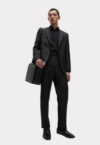 Zwarte suit met een getailleerde pasvorm, gecombineerd met een overhemd met knopen. Vlotte textuur, slank ontwerp en gepolijste zwarte schoenen. Inclusief een zwarte schoudertas.