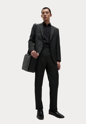 Abito nero dalla vestibilità su misura, abbinato a una camicia con bottoni. Texture liscia, design elegante e scarpe nere lucide. Include una borsa a tracolla nera.