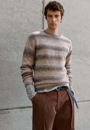 Joven con un suéter beige a rayas, camisa gris de manga larga debajo, pantalones marrones y cinturón negro, posando junto a una pared de hormigón.