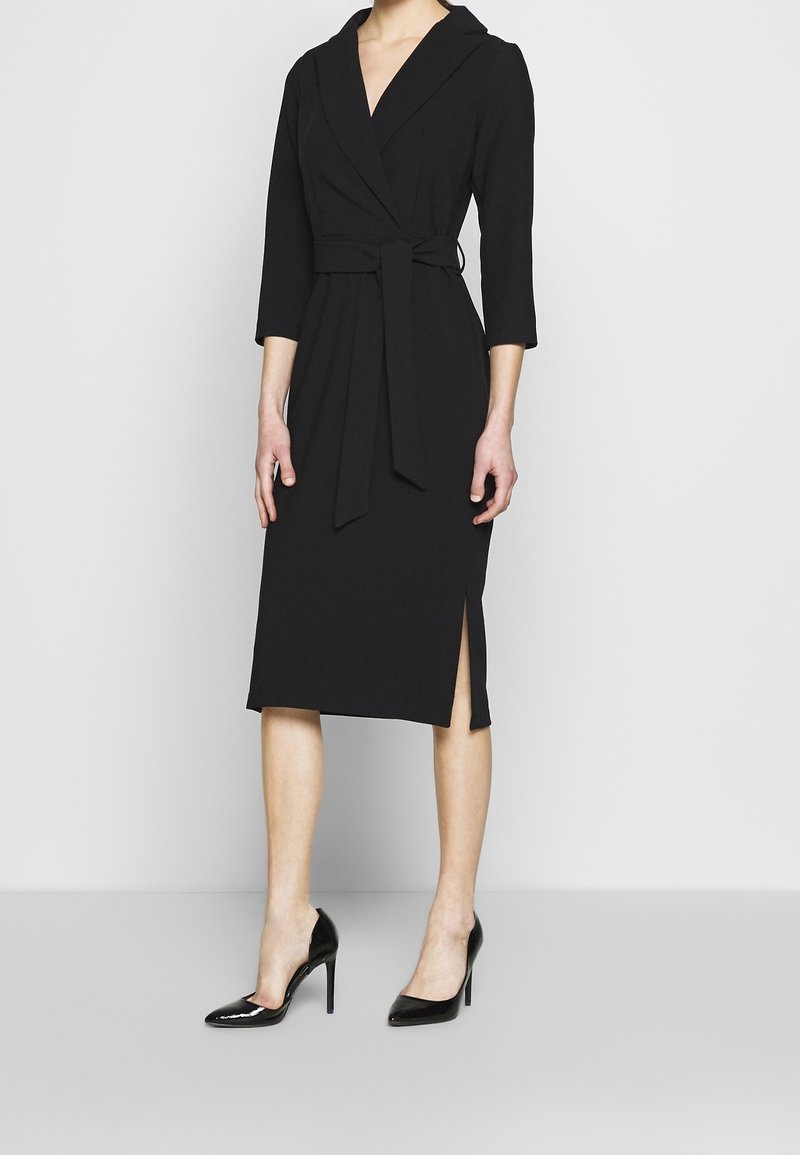 Femme portant une robe portefeuille noire longueur genou avec manches trois-quarts et ceinture, associée à des chaussures à talons noirs.