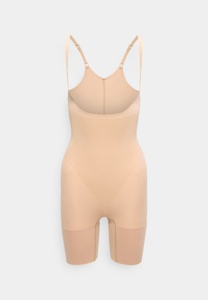 Bež oblikujuća odjeća za cijelo tijelo s podesivim remenima na ramenima i nogavicama do bedara, prikazana na jednostavnoj svijetloj pozadini.