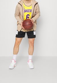 Basketbalista drží míč, má na sobě žlutý dres s fialovými akcenty, béžovou mikinu na zip, černé šortky s bílými záplatami a bílé boty.