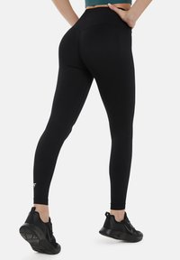 Črne legice z visokim pasom, narejene iz raztegljivega materiala z gladko teksturo in diskretnim logotipom blizu gležnja. Oblečene z črnimi športnimi čevlji.