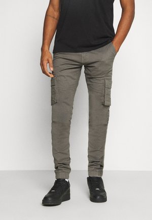 Męskie spodnie cargo w oliwkowym zielonym kolorze z bocznymi kieszeniami i elastycznymi mankietami, w połączeniu z czarnymi trampkami i czarną koszulką, stojące na prostym tle.