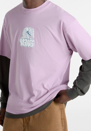 Homme portant un T-shirt lilas par-dessus une chemise à manches longues sombre, pantalon beige, avec un graphisme de tête de serpent blanc et un texte stylisé sur le devant du T-shirt.