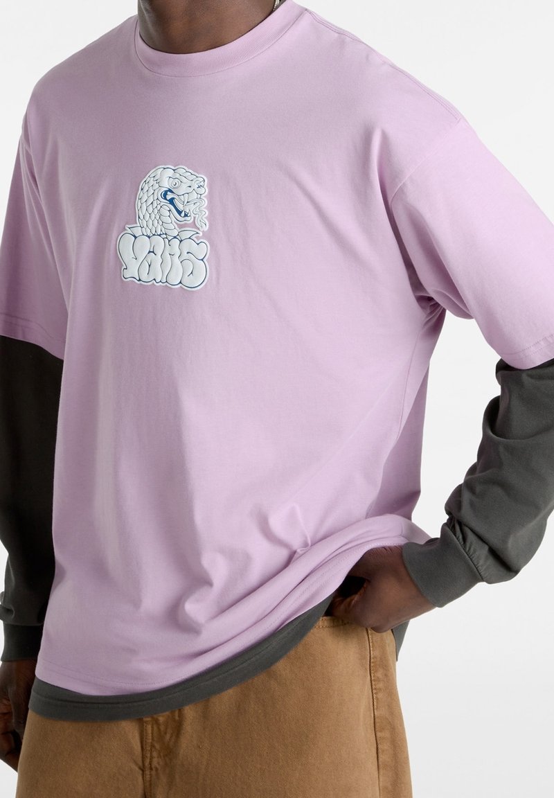 Homme portant un T-shirt lilas par-dessus une chemise à manches longues sombre, pantalon beige, avec un graphisme de tête de serpent blanc et un texte stylisé sur le devant du T-shirt.