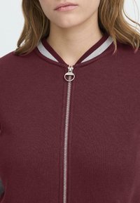 Veste zippée bordeaux avec une fermeture éclair argentée et un col rayé gris. Matériau doux avec une texture lisse, présentant un design ajusté.