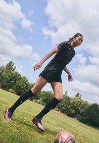 Crna sportska odjeća s majicom kratkih rukava i kratkim hlačama, uparena s crnim čarapa do koljena i šarenim nogometnim kopačkama, na travnatom terenu.