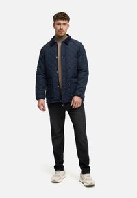Chaqueta de napa azul marino con cuello, cierre de botones a presión y dos bolsillos frontales, llevada sobre un suéter beige y jeans oscuros.