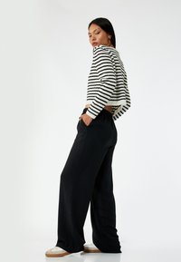 Beskåret stribet sweater i sort og hvid, parret med løse sorte bukser med elastisk talje. Modellen står med hænderne i lommerne.