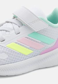Chaussure de sport blanche avec tige en mesh, mettant en avant des accents pastel en menthe, rose, jaune et violet, et une sangle ajustable pour un maintien sécurisé.