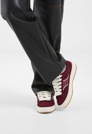 Jambes croisées portant un pantalon noir à jambes larges et des baskets bordeaux avec des lacets et semelles blancs sur un fond blanc.