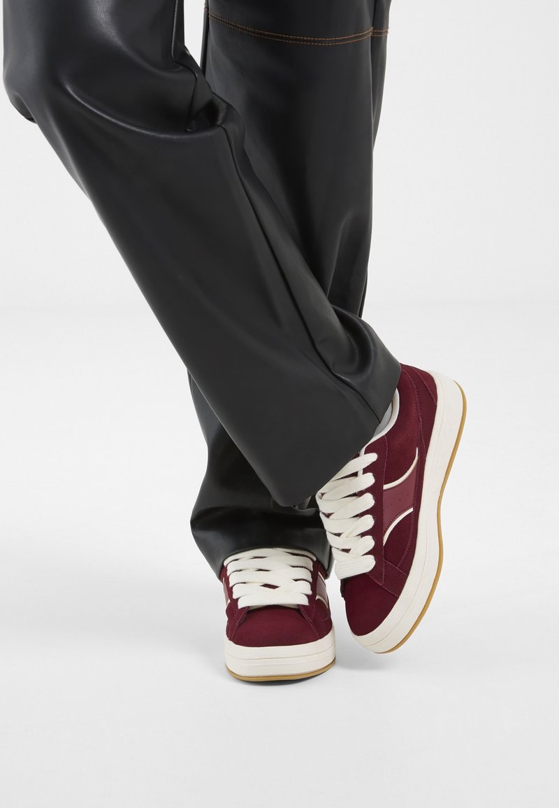 Jambes croisées portant un pantalon noir à jambes larges et des baskets bordeaux avec des lacets et semelles blancs sur un fond blanc.