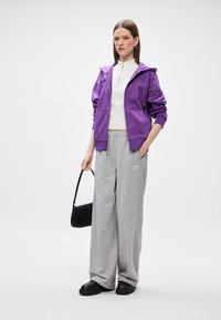 Fialová mikina na zips cez biely top s vysokým golierom, skombinovaná s voľnými sivými teplákmi. Čierna kabelka a čierne topánky dotvárajú outfit.