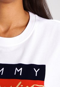 Bílý bavlněný svetr s kulatým výstřihem a grafickým potiskem, s odvážným písmem v námořnické modré a červené barvě přes hruď.