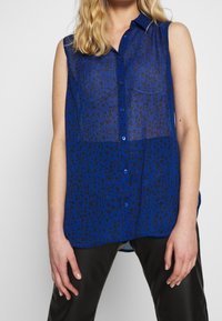 Blusa sin mangas azul de tela translúcida con un estampado de leopardo negro, con botones en la parte delantera y un cuello, combinada con pantalones negros.