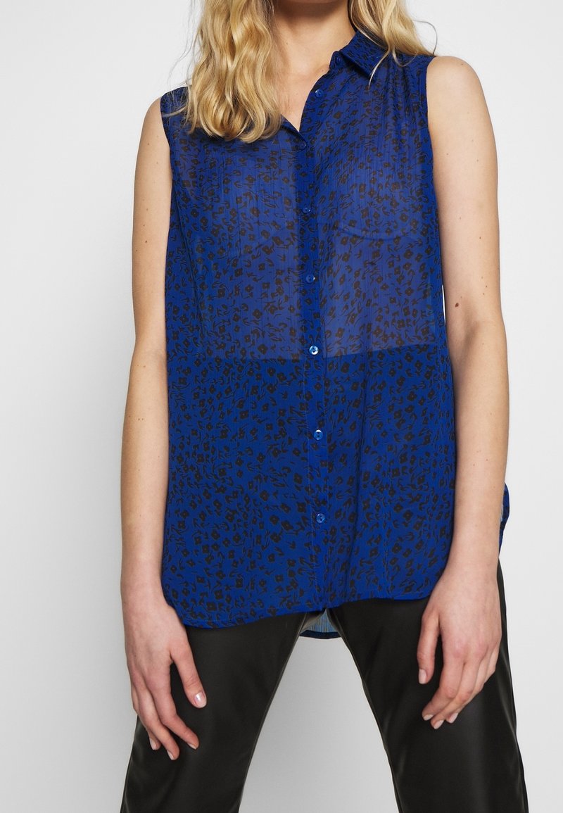 Blusa sin mangas azul de tela translúcida con un estampado de leopardo negro, con botones en la parte delantera y un cuello, combinada con pantalones negros.