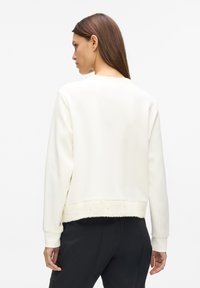 Biela mikina s textúrovaným krémovým lemom, dlhými rukávmi a voľným strihom. Dizajn dopĺňa hladká látka a rebrované manžety.