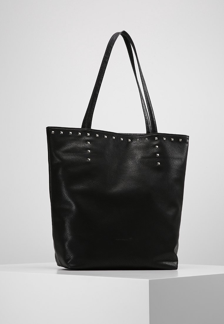 Sac fourre-tout en cuir noir avec double anse, décoré de petits clous carrés métalliques le long du bord supérieur, présenté sur un socle blanc.