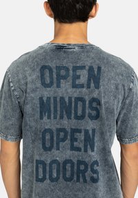Persoon met kort, donker haar die een vervaagde grijze t-shirt draagt met de tekst "OPEN MINDS OPEN DOORS" op de achterkant gedrukt.