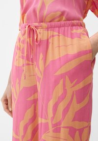 Losse broek in roze stof met grote oranje bladpatronen. Heeft een elastische tailleband met een koord en zijzakken.