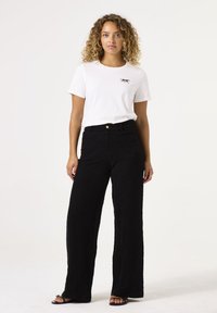 Camiseta blanca con un pequeño gráfico negro en el pecho, combinada con pantalones anchos negros texturizados y sandalias negras. Fondo plano y limpio.