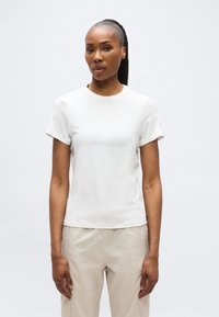 Γυναίκα με μακριά πλεξούδα αλογοουρά, φοράει απλό λευκό κοντομάνικο μπλουζάκι και μπεζ παντελόνι, στέκεται μπροστά σε απλό φωτεινό φόντο.