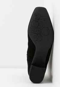 Suola di una scarpa elegante nera con motivi impressi che presentano monete e la parola "KAISER" su una superficie bianca.