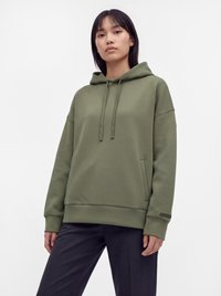 Mujer que lleva una sudadera con capucha verde oliva con cordones y pantalones oscuros, de pie frente a un fondo blanco liso.