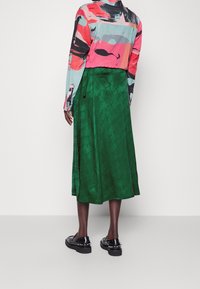 Falda midi de satén verde con un patrón sutil, combinada con una blusa impresa colorida. Cuenta con un cierre de cremallera y zapatos negros con suela gruesa.