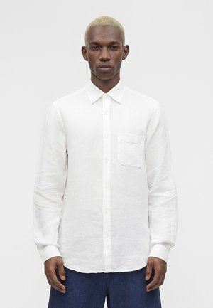 Mand med kort blondt hår iført en hvid knappet langærmet skjorte og mørkeblå bukser, vendt mod kameraet foran en ensfarvet baggrund.