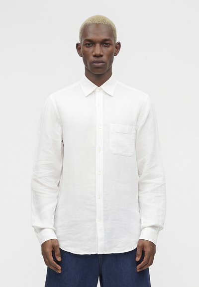 Homme aux cheveux blonds courts portant une chemise blanche à manches longues boutonnée et un pantalon bleu foncé, face à un fond uni.