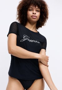 Fekete, rövid ujjú póló áttetsző hálós díszítéssel, elöl strasszkövekkel kiemelt "Guess" felirattal. Simaság, laza dizájn.