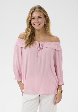 Jeune femme portant un haut rose à épaules dénudées avec un détail noué et un pantalon blanc, souriante devant un fond clair uni.